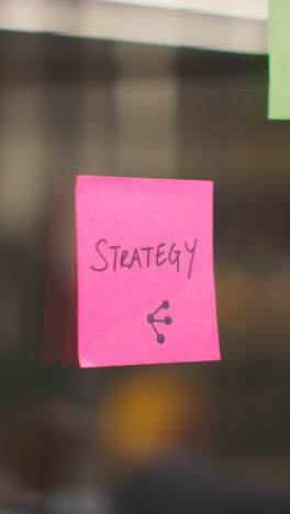 Vertical-Video-Close-Up-Of-Woman-Putting-Sticky-Note-With-Strategy-Written-On-It-Onto-Transparent-Screen-In-Office